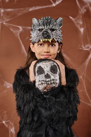 Porträt eines lächelnden Mädchens in Wolfsmaske und schwarzer Kleidung, das gruseliges Spielzeug umarmt, Halloween, Nahaufnahme