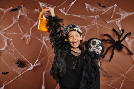 cheerful preteen girl yaying and holding scary toy and bucket of sweets, Halloween concept Poster 676677298