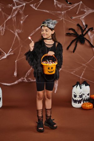 Foto de Feliz niña preadolescente mirando piruleta y sosteniendo cubo de dulces, concepto de Halloween - Imagen libre de derechos