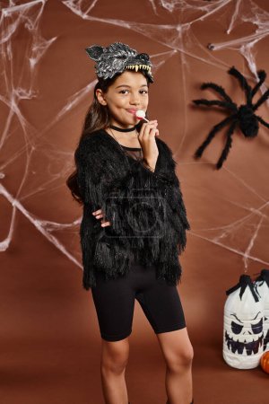 niña preadolescente con brazos cruzados y piruleta sobre fondo marrón con tela de araña, concepto de Halloween