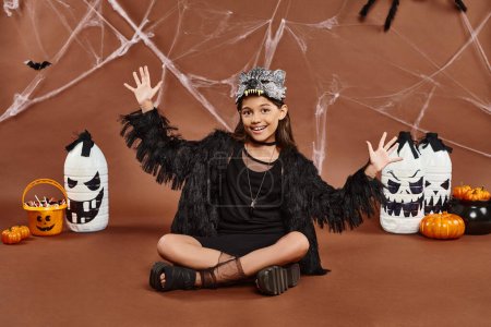 chica feliz en máscara de lobo y traje negro sentado con las piernas cruzadas y mostrando las palmas abiertas, Halloween