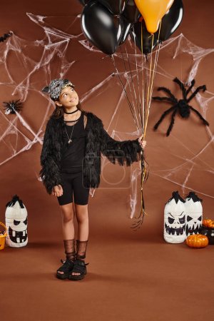 smiley girl holds black and orange balloons on brown backdrop with web and spiders, Halloween