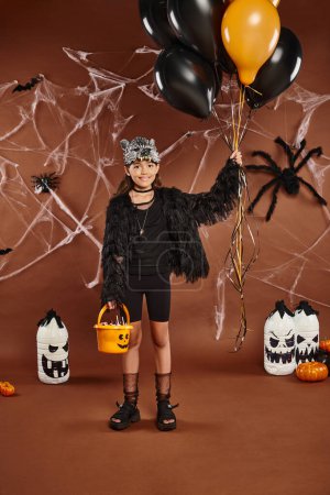 Smiley-Mädchen hält schwarze und orangefarbene Luftballons und Eimer mit Süßigkeiten, Halloween-Konzept