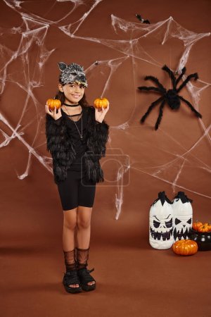 preteen girl standing in wolf mask holds pumpkins on brown backdrop, Halloween concept