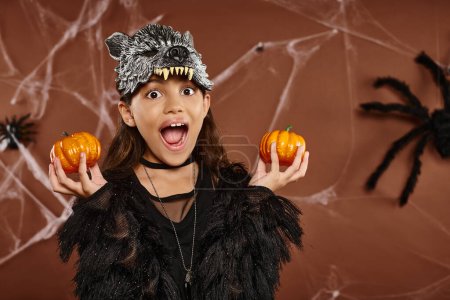 close up excited girl in wolf mask holds pumpkins in her hands with spider on backdrop, Halloween