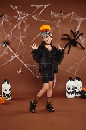 preteen girl holding pumpkin on her head with raised hands, brown backdrop with web, Halloween magic mug #676678180