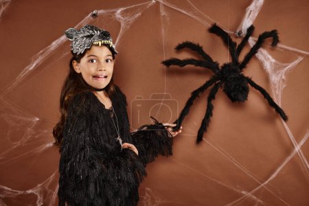 Téléchargez les photos : Gros plan fille effrayée dans le masque de loup et costume noir touchant araignée sur fond brun, Halloween - en image libre de droit