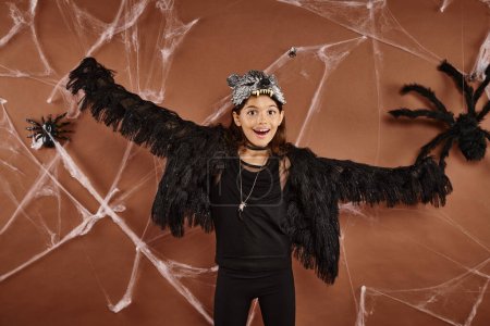 glückliches Mädchen mit geöffneten Armen in schwarzer Kunstpelztracht mit Spinnennetz auf braunem Hintergrund, Halloween
