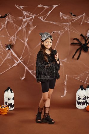 alegre niña preadolescente de pie todavía en traje de piel sintética negro con fondo marrón, Halloween