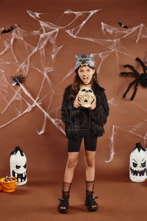 niña preadolescente haciendo muecas y sosteniendo el cráneo en el fondo marrón con telaraña y linternas, Halloween