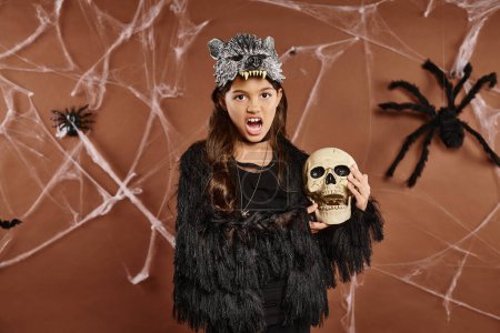 cerrar chica sorprendida con la boca abierta sostiene cráneo con fondo marrón, concepto de Halloween