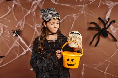 niña preadolescente sonriente sosteniendo cráneo y cubo de dulces, fondo marrón con telaraña, Halloween
