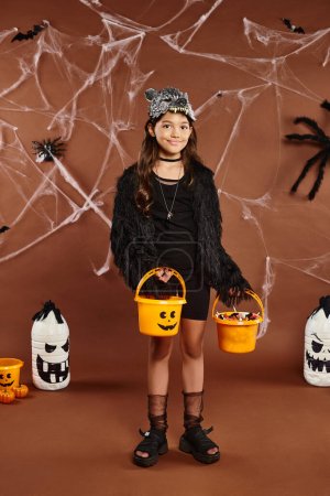 cheerful kid in wolf mask holds two buckets of sweets with bats and spiders on backdrop, Halloween
