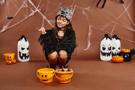 preteen girl squats down near buckets of sweets and holds candy, Halloween concept