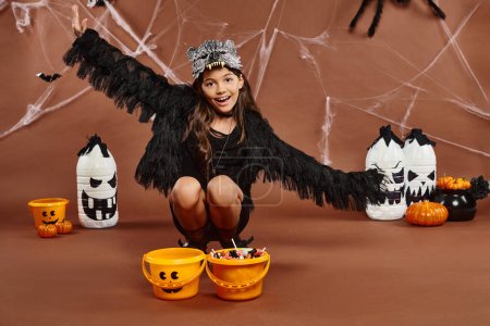 cheerful kid in wolf mask squats down near pumpkin buckets with open arms, Halloween