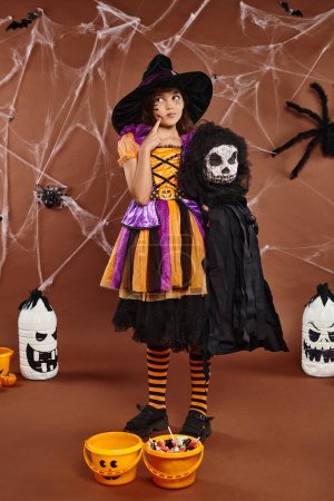 preteen girl in witch hat with spooky toy and touching her cheek and looks away, Halloween