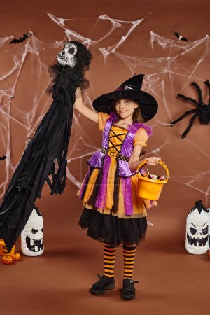 happy kid in witch hat with spiderweb makeup holding bucket of sweets and spooky toy, Halloween