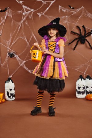 cute preteen in witch hat holds bucket of sweets on brown backdrop with spiderwebs, Halloween