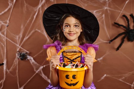 happy girl in witch hat with spiderweb makeup holding bucket of sweets on brown with spider net