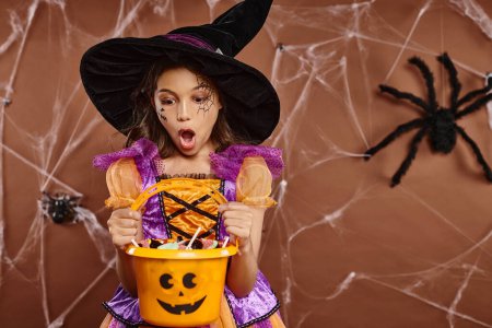 Schockiertes Mädchen mit Hexenhut und Spinnennetz-Make-up beim Anblick von Süßigkeiten im Halloween-Eimer auf braun