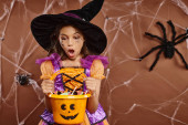 shocked girl in witch hat with spiderweb makeup looking at sweets in Halloween bucket on brown Stickers #676679570