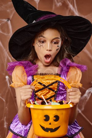 shocked kid in witch hat and Halloween costume looking at sweets in bucket on brown background