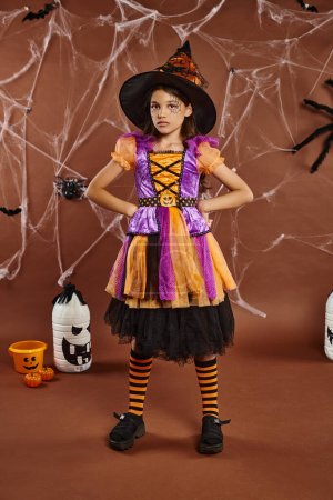 serious girl in Halloween costume and witch hat standing with hands on hips on brown backdrop