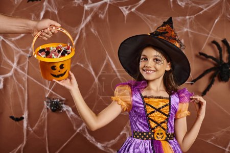 happy girl in witch hat and Halloween costume looking at camera near hand holding sweets in bucket
