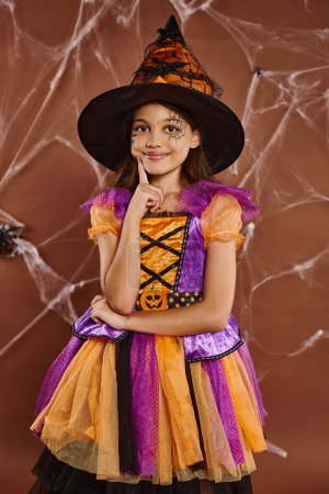 glückliches Mädchen mit Hexenhut und Halloween-Kostüm in der Nähe von Spinnweben auf braunem Hintergrund, gruselige Jahreszeit