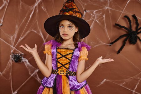 Verwirrtes Mädchen mit Hexenhut und Halloween-Kostüm in der Nähe von Spinnweben auf braunem Hintergrund, gruselige Jahreszeit