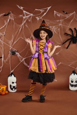 Photo for Happy girl in witch hat and Halloween costume gesturing near cobwebs on brown, spooky season - Royalty Free Image