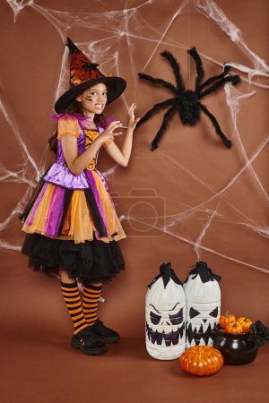 Gruseliges Mädchen mit Hexenhut und Halloween-Kostüm knurrt in der Nähe einer falschen Spinne auf braunem Hintergrund