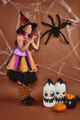 spooky girl in witch hat and Halloween costume growling near fake spider on brown background Stickers #676680240