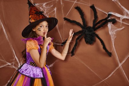 Gruseliges Mädchen mit Hexenhut und Halloween-Kostüm, das Schweigen in der Nähe einer falschen Spinne auf braunem Hintergrund zeigt