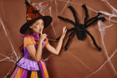 spooky girl in witch hat and Halloween costume showing hush near fake spider on brown background magic mug #676680280