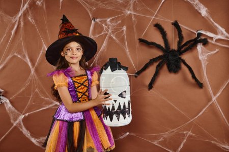 happy girl in witch hat and Halloween costume standing with spooky decor on brown background