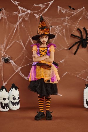 sad girl in witch hat and Halloween costume standing with folded arms on brown backdrop, cobwebs