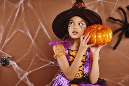 spooky girl in witch hat and Halloween costume standing with pumpkin on brown backdrop, cobwebs