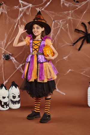 joyful girl in witch hat and Halloween costume standing with pumpkin and waving hand on brown