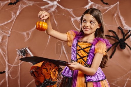 positive child in Halloween witch costume holding pumpkin near pointed hat on brown background