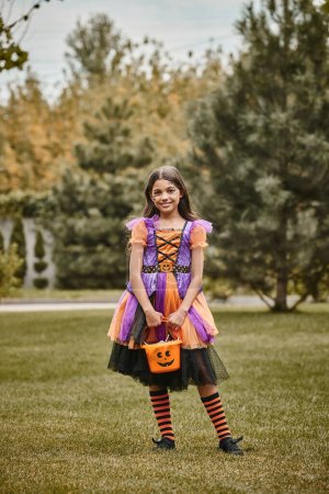 niedliches Kind im Halloween-Kostüm, das mit einem Eimer süßer Bonbons auf grünem Gras steht, Mädchen im Kleid
