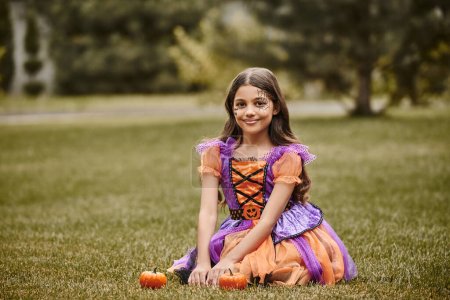 cheerful girl in Halloween costume sitting in vibrant dress near to tiny pumpkins on green grass
