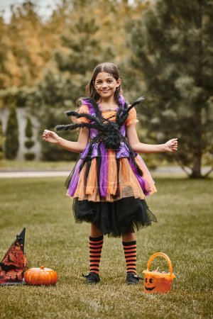happy girl in Halloween costume with spider standing near pumpkin, pointed hat and candy bucket