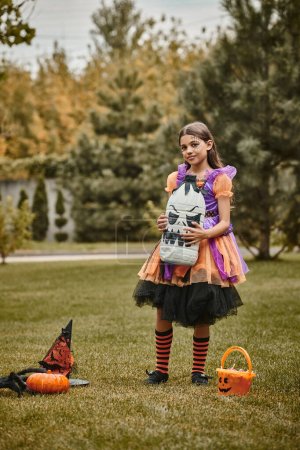 Téléchargez les photos : Fille en costume d'Halloween tenant la décoration effrayante bricolage près de la citrouille, chapeau pointu et seau de bonbons - en image libre de droit