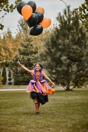 fröhliches Mädchen im Halloween-Kostüm mit Luftballons und Süßigkeiteneimer beim Laufen auf grünem Gras