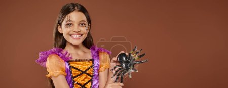 joyous girl in Halloween costume with spiderweb makeup holding fake spider on brown backdrop, banner