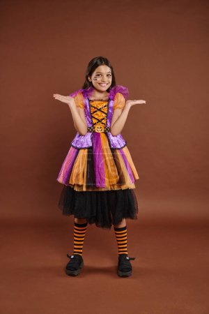 funny girl in Halloween costume with spiderweb makeup smiling and gesturing on brown backdrop