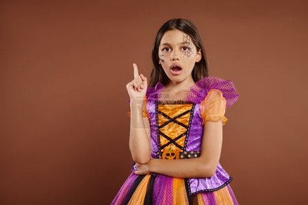 Photo for Amazed girl in Halloween costume with spiderweb makeup having idea, pointing up on brown backdrop - Royalty Free Image