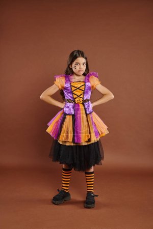 missmutiges Mädchen im Halloween-Kostüm, das mit den Händen auf Hüften vor braunem Hintergrund steht, 31. Oktober