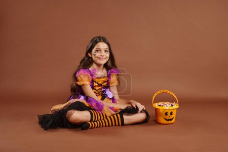 fröhliches Mädchen im bunten Halloween-Kostüm sitzt neben Eimer mit Bonbons auf braunem Hintergrund